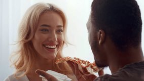 A biracial couple shares a joyful moment at home, playfully feeding each other pizza. Their affection shines through as they embrace love and connection during a casual meal. - Powered by Shutterstock - Get 15% off with code: PIKWIZARD15