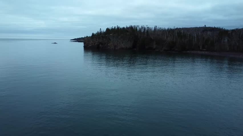Aerial view of dramatic rocky cliff lined with bare trees, reflected in the serene waters of a quiet lake under, video shot in Minnesota.
