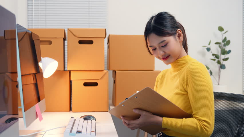 Asian woman checking inventory and writing on clipboard, surrounded by cardboard boxes in her office