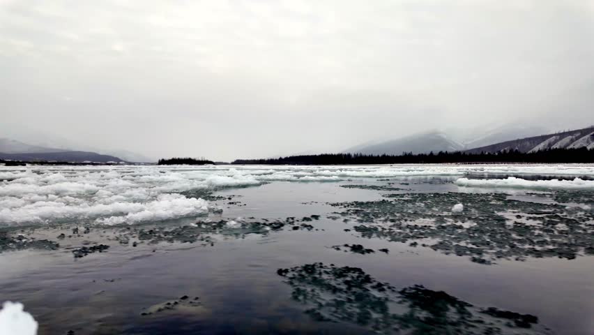 Rugged ice formations dot a tranquil river surrounded by snow-covered mountains. The calm water reflects the overcast sky, showcasing a serene winter atmosphere.