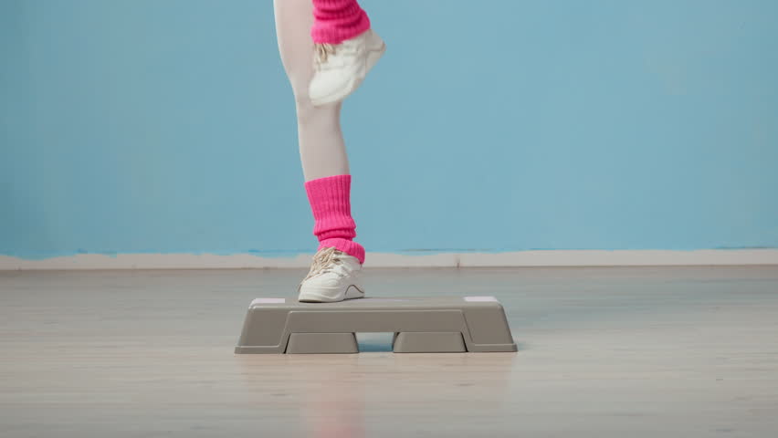 Wide tilt up shot of female millennial in retro pink and purple activewear practicing aerobic steps on platform in studio with pastel blue walls