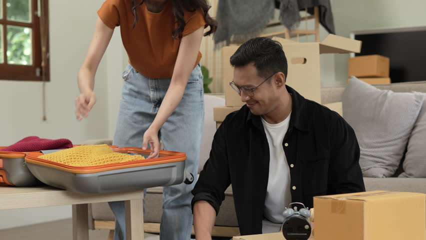 A happy young couple sits on a sofa, packing belongings into boxes, preparing to move to a new house, enjoying teamwork, organizing items, showing excitement, and embracing a fresh start