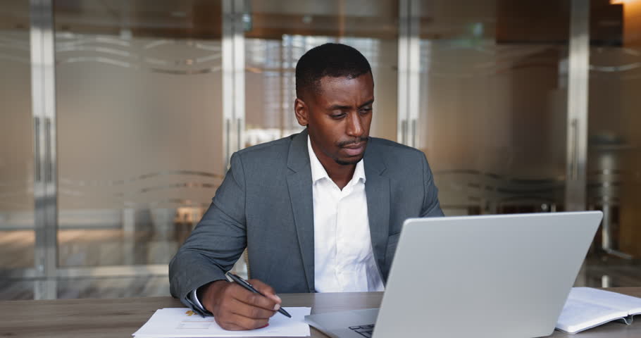 Serious African businessman dressed suit working at desk in office, researching information, making notes on paper and using laptop, check reports, websurfing info on internet, jotting important data