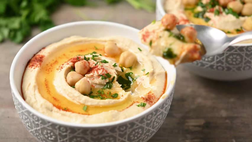 person eating with a spoon chickpea hummus with olive oil, parsley and paprika in bowl on wooden table