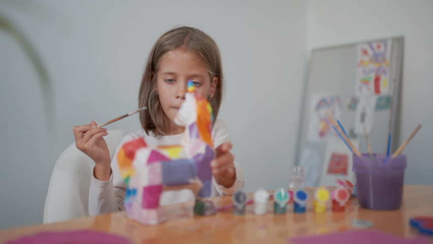 School-aged girl with blonde hair sitting at table at home painting plaster figurines using glue and colorful gouache paints engaging in creative arts and crafts activity for child development 