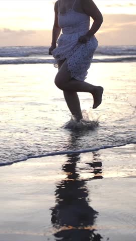 Silhouette of a beautiful girl in a white dress running along the ocean coastline along the foam of water at sunset. Happy ending of vacation. Vertical shot.