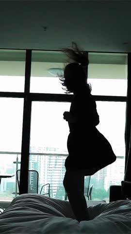Happy woman jumping on bed in luxury apartment, super slow motion, 250fps. Backlight. Cool hotel room with huge window bg Vertical Shot.