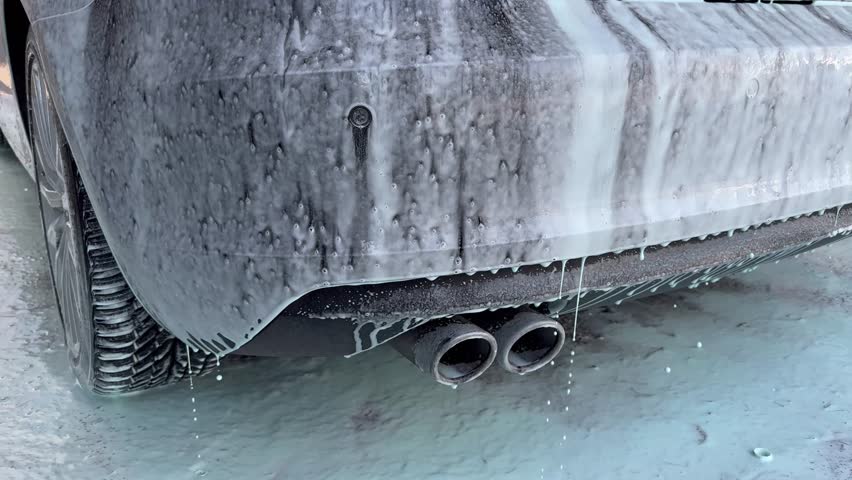 The rear of a car covered in soap suds during a wash, featuring dual exhaust pipes and water dripping down the soapy surface
