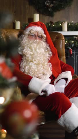 Santa Claus on xmas eve sitting in armchair pulls out old red silk album near green christmas tree with toys and lights. Wide shot slider dolly movement Vertical Shot.