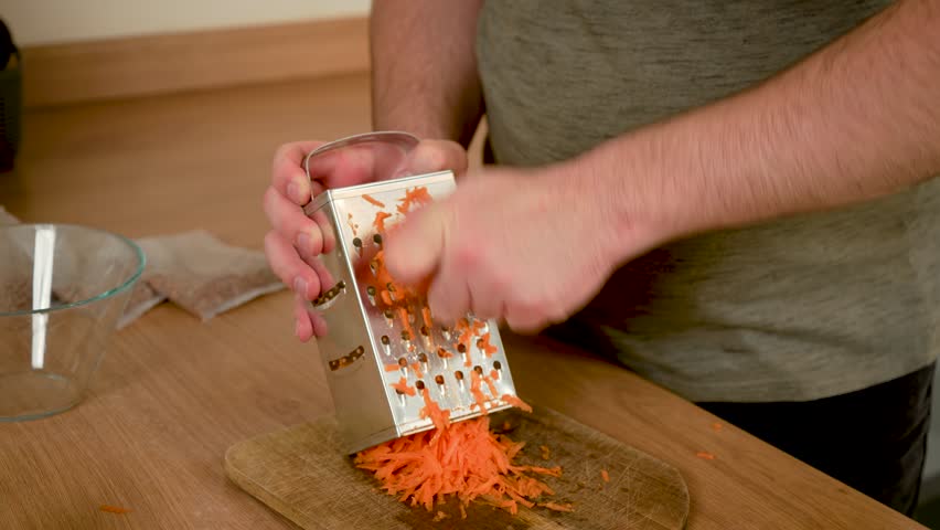 Man grating carrot onto wooden board. Male shredding orange vegetable with kitchen grater. Guy working with box grater to create shredded carrot. Gentleman preparing grated carrot using grater