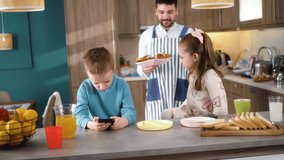 Two little kids brother and sister playing games or watching something on smartphones at the table while their dad prepares breakfast for them in the kitchen - Powered by Shutterstock - Get 15% off with code: PIKWIZARD15