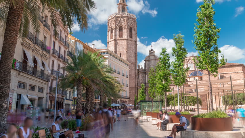 Panoramic timelapse of Plaza de la Reina in Valencia, Spain. Lively square with a historic church tower, bustling cafes and a central fountain under the warm Mediterranean cloudy sky.
