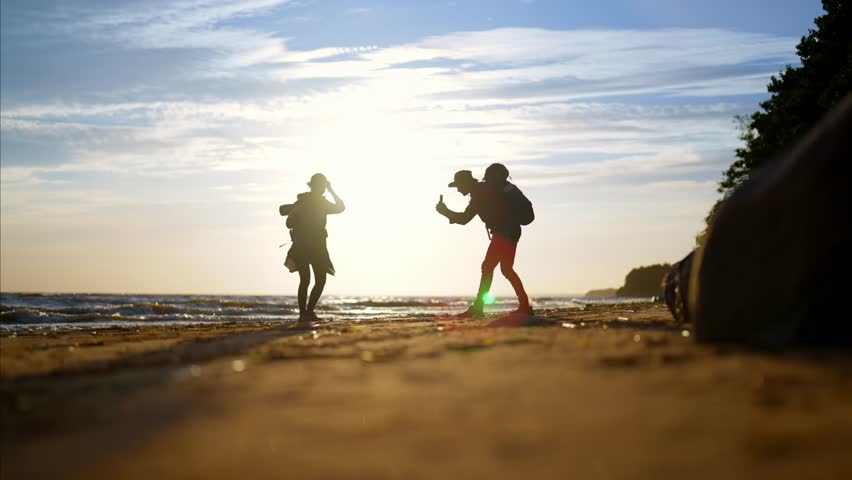 Cinematic general shot with hikers in nature, taking pictures by phone on beach. Silhouette of travelers against bright sun in blue sky, woman posing for camera of smartphone, mobile photo shooting