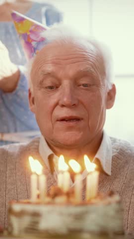 Old man celebrating his birthday, blowing burning candles on cake, closeup portrait, vertical video. Funny grandfather with birthday cap feeling joy and happiness, family gathering at home, happy fest