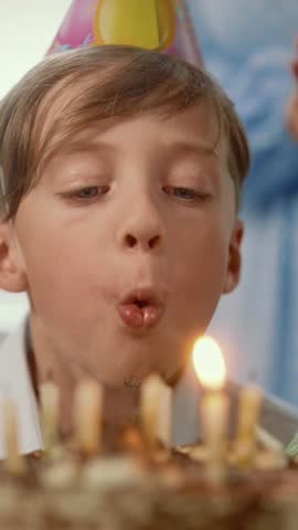 Joyful boy with birthday cap on head blowing flame of candles on cake, closeup view, vertical portrait. Happy childhood moments, family celebration birthday of beloved son, grandchild and brother