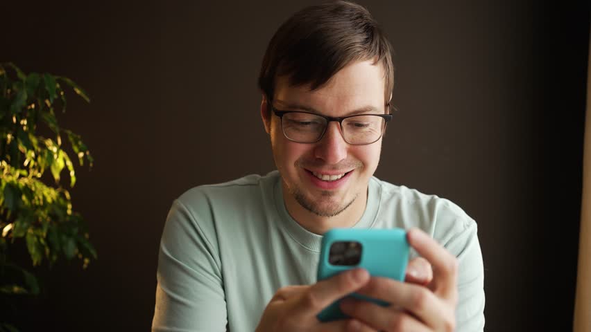 Young adult man wearing glasses and casual clothes is using smartphone, browsing internet and social media, smiling and having fun on dark background with houseplant