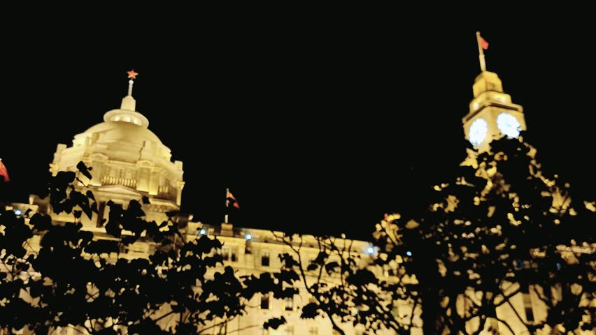 Tourist take phone photo at night of large building with two clocks on top of it in Shanghai, China. The building is lit up at night