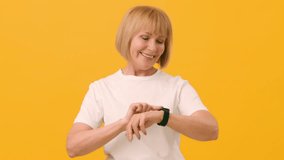 A cheerful older woman looks at her smartwatch with a smile while standing in front of a bright yellow background. She enjoys the moment, showcasing her engagement with technology. - Powered by Shutterstock - Get 15% off with code: PIKWIZARD15