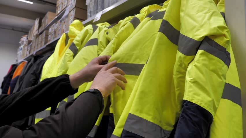 A person inspects the Rows of  safety jackets organized by size, are neatly arranged, showcasing their reflective stripes and vibrant color, ideal for visibility and protection at work.
