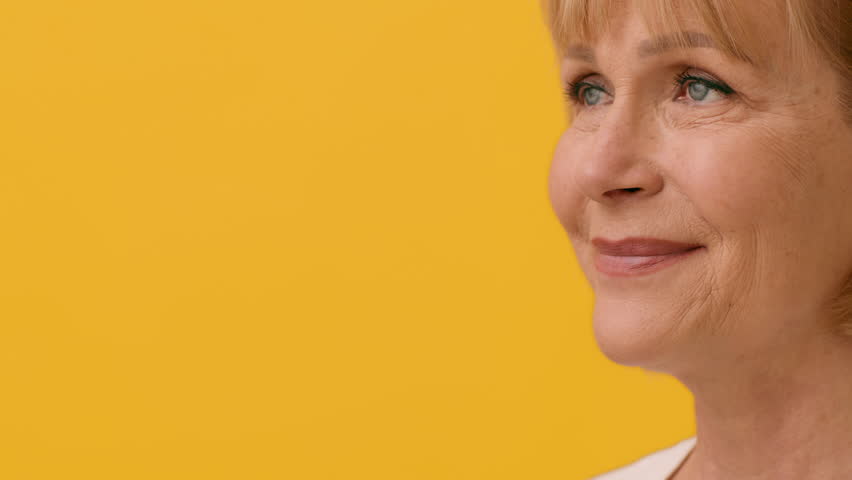 A smiling older woman with short blonde hair stands in front of a vibrant yellow background, showcasing a warm and cheerful demeanor. The sunlight highlights her happy expression, copy space