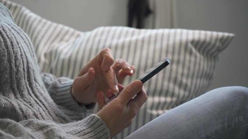Old woman browsing news on her smartphone, sitting by a window and engaged in digital reading