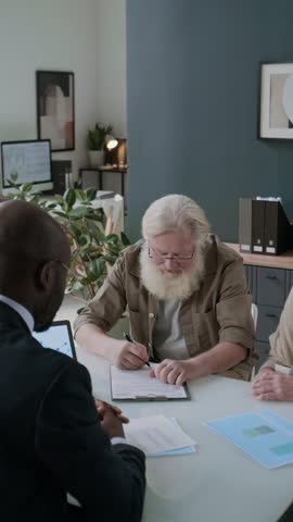 Vertical shot of elderly man sitting next to his wife at office desk, signing contract and giving handshake to financial advisor