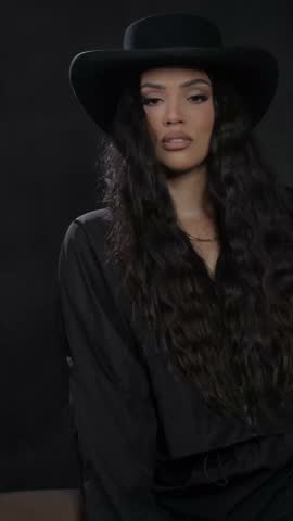 A brunette with wavy hair in a black cowboy hat and a golden chain around her neck poses for a photographer
