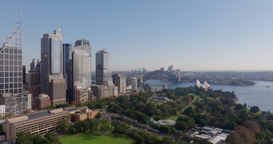 Aerial shot of Sydney CBD skyline during the day. Drone flying towards botanic gardens, harbour bridge, opera house and Sydney Harbour with traffic and clear blue sky visible