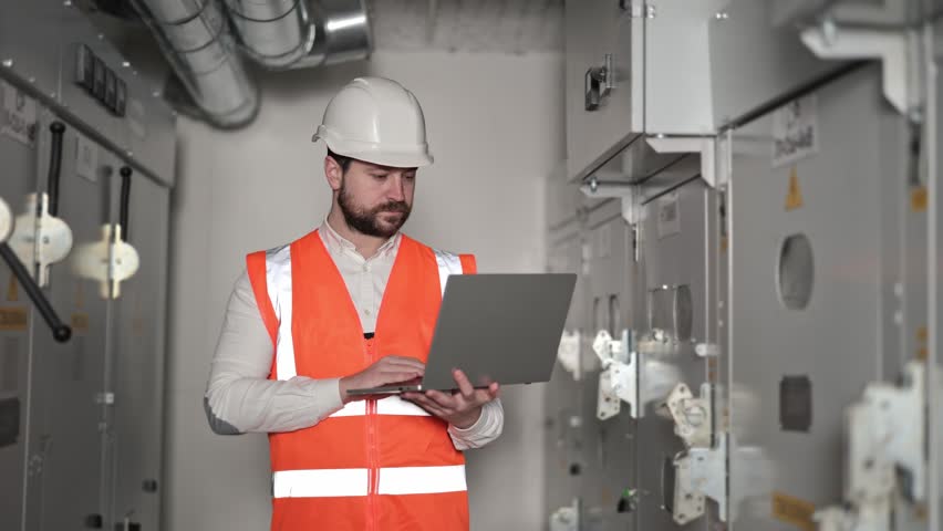 Serious electrical engineer with laptop inspects switchboard. Bearded man in orange vest and white hardhat types on laptop keyboard. Industrial environment with electrical control panels and wiring.