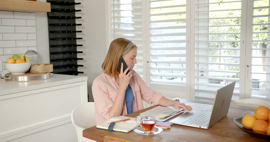 Working from home, woman using laptop and talking on smartphone in kitchen. Remote working, telecommuting, home office, multitasking, technology, communication, unaltered,