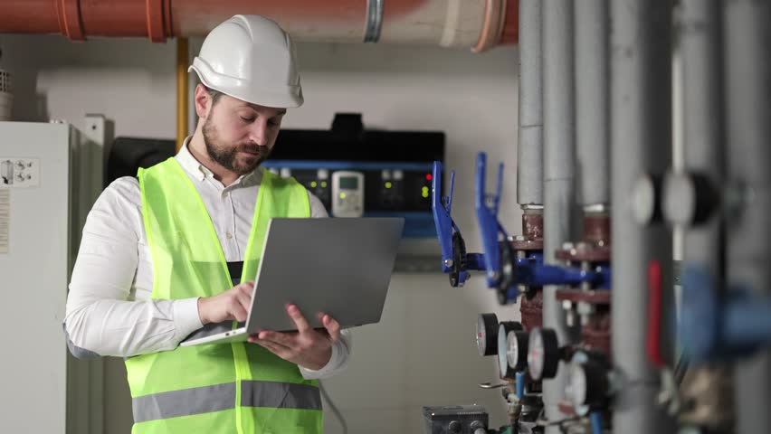 Smiling engineer with laptop in boiler room. Industrial worker with laptop. Professional worker in heating system equipment. Control of heating system in apartment building.