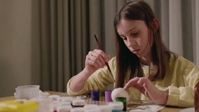 A teenage girl concentrating on painting an Easter egg with bright colors, surrounded by paints and brushes at a festive table. High quality 4k footage - Powered by Shutterstock - Get 15% off with code: PIKWIZARD15