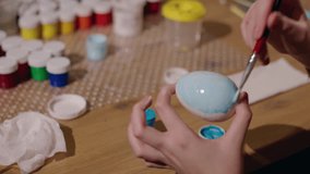 Close-up of hands painting an Easter egg with light blue and white colors, surrounded by brushes and paint jars on a wooden table. High quality 4k footage - Powered by Shutterstock - Get 15% off with code: PIKWIZARD15