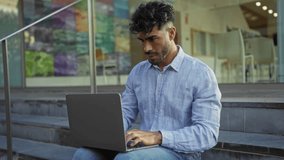Young man with laptop sitting outside on urban steps, smiling and working, wearing casual blue shirt in city environment, engaged in video call. - Powered by Shutterstock - Get 15% off with code: PIKWIZARD15