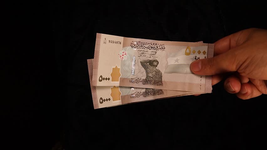 Hand holding a bundle of 5000 Syrian pound banknotes passes it to another hand in a seamless transaction. Represents Syrian currency exchange, financial transfer, business dealings in a clean setting.