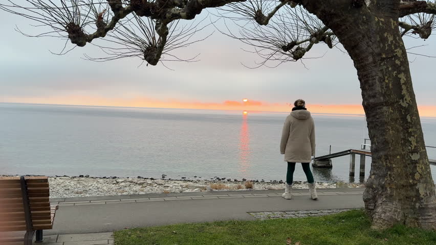 A woman in a beige coat stands near a tree and paved walkway, gazing at the sunrise over Lake Constance (Bodensee). The calm water reflects the warm glow of the rising sun. Filmed in Germany