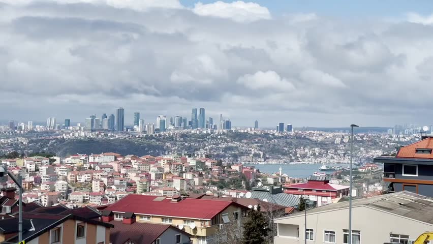 Panoramic view of Istanbul from a high vantage point. The foreground features low-rise buildings, while the background showcases a cityscape with skyscrapers. Sunlight breaks through