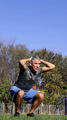 Middle-Aged Man with Gray Hair Performing Squats on a Sports, Grass Field. Background includes blue sky, green trees, and various sport fields. Ideal for promoting fitness and a healthy life style.