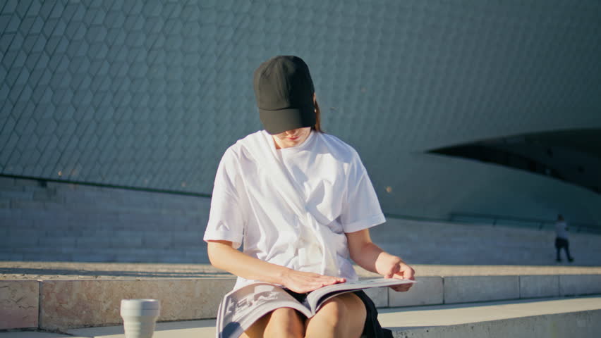 Trendy woman reading magazine sitting urban stairs summer day closeup. Relaxed lady in black cap flipping pages fashion journal relaxing on street. Young brunette looking articles at sunny square.