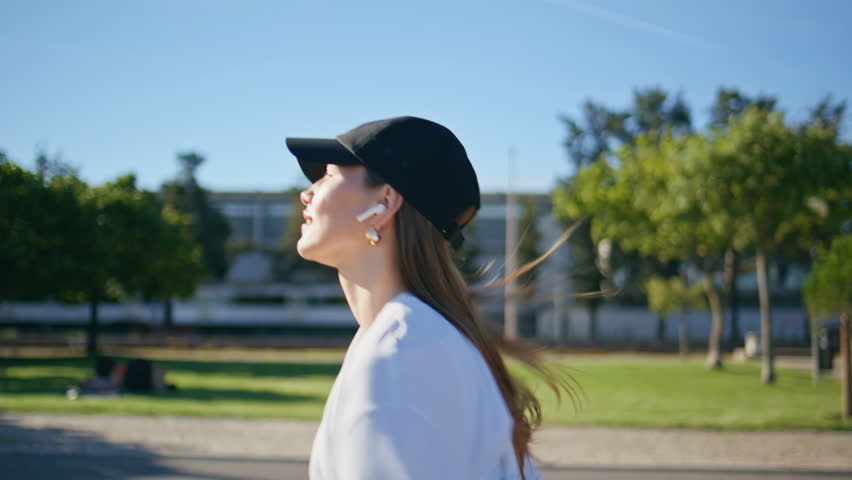 Smiling girl running city sunny road listening music in wireless earphones closeup. Happy female runner enjoying sport activity at summer street. Energetic woman jogging green urban area in black cap.