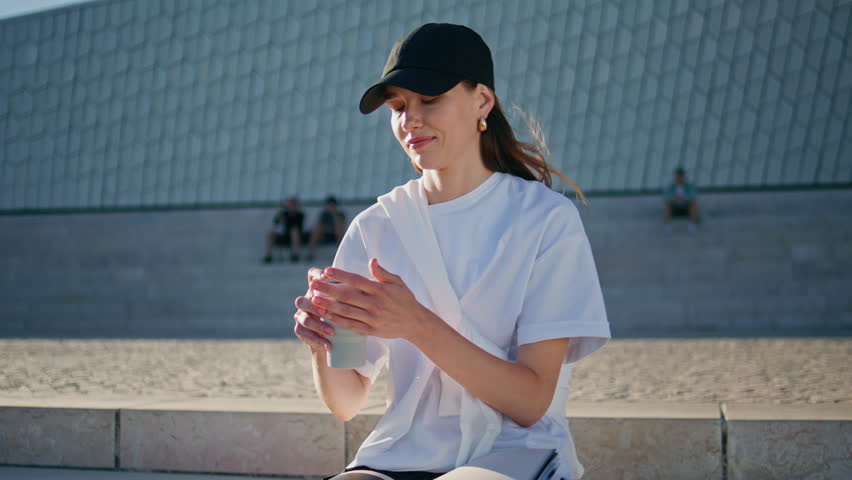 Serene woman drinking beverage enjoying morning sunlight outdoors closeup. Relaxed girl in cap sipping hot coffee feeling calmness on sunny street. Beautiful lady relaxing sitting urban stairs alone.