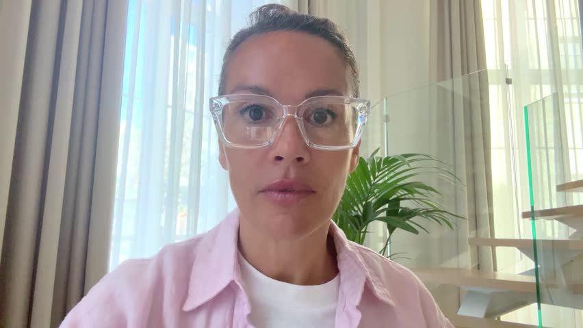 Professional woman with tied hair and glasses sitting at her home workspace, working on her tablet in a focused manner. Woman viewed from camera phone touching screen alone at home with serious face