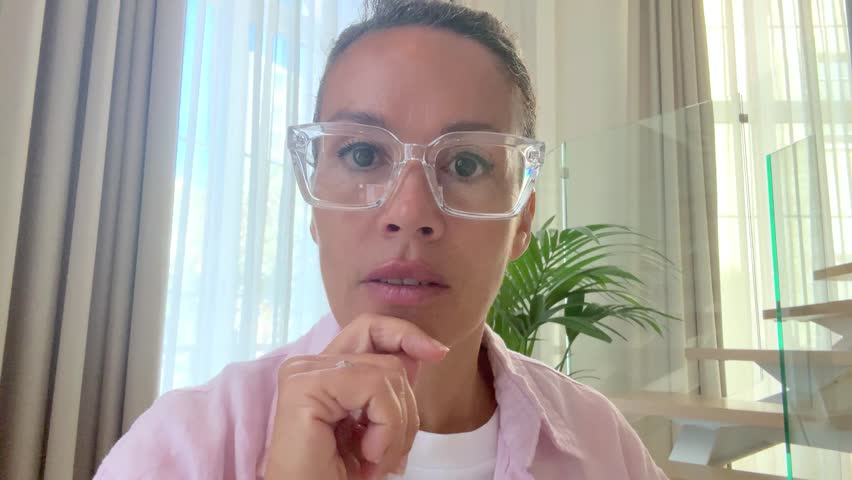 Woman viewed from camera phone touching screen alone at home with serious face. Professional woman with tied hair and glasses sitting at her home workspace, working on her tablet in a focused manner.