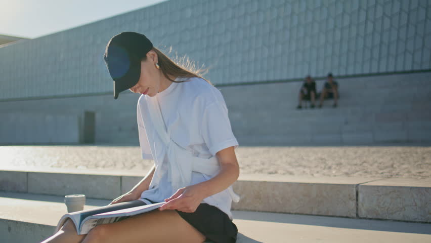 Relaxed woman flipping pages magazine sitting sunny street with coffee cup. Carefree young lady in black cap reading fashion journal on urban stairs. Calm brunette enjoying summer day leisure outdoors