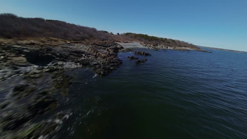 FPV drone shot speeds along rugged coastline of New Hampshire, USA, flying just above water and over rocks lining shore.