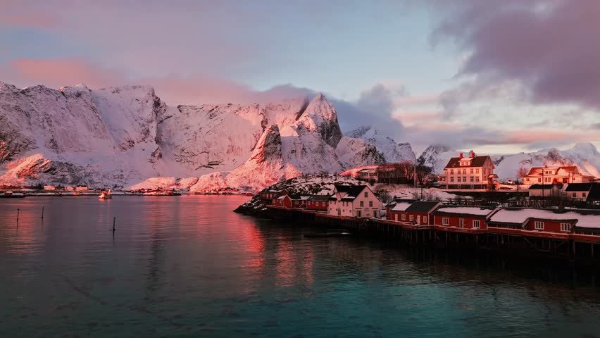 Flying over bright calm waters at Sakrisoy, Lofoten towards the sunrise with snow-covered mountains in the morning sunshine