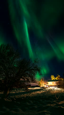 Vertical, aurora borealis glowing green pillars over cozy winter cabin surrounded by snow in Finland