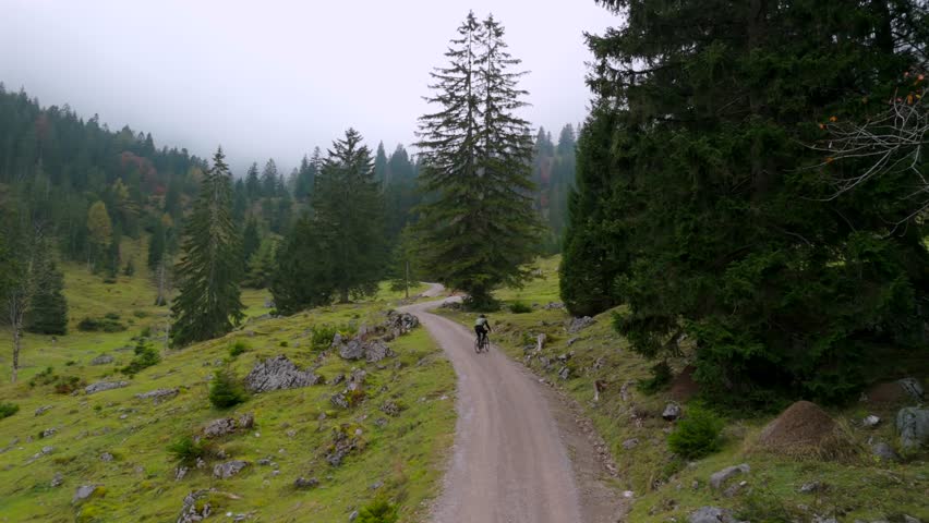 A male cyclist on a gravel bike rides uphill on a mountain road through a foggy autumn forest in Germany. Drone footage captures him cycling through the misty atmosphere of the Alps
