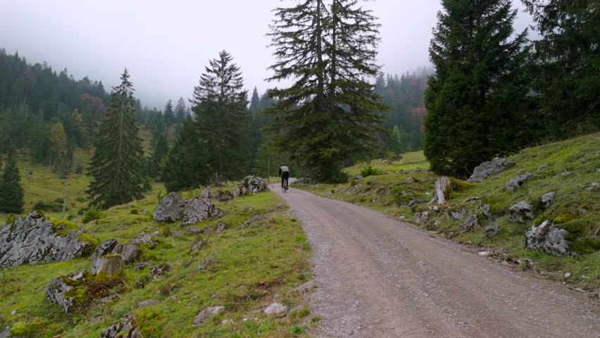 A male cyclist on a gravel bike rides uphill on a mountain road through a foggy autumn forest in Germany. Drone footage captures him cycling through the misty atmosphere of the Alps
