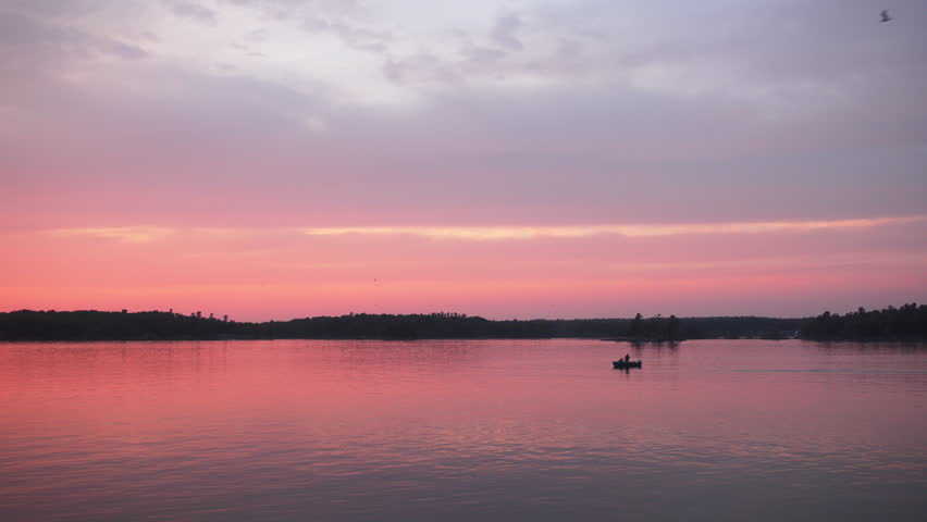 Handheld shot of a lone fisherman in a boat silhouetted against a vibrant pink and orange sunset on the St. Lawrence River at Wellesley Island State Park, NY.